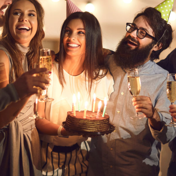 Three friends celebrating a birthday party while wearing party hats. Most are holding champagne but the one in the center is holding a lit birthday cake.
