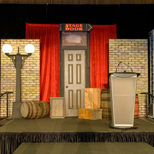 The stage at the 2023 Laughs + Libations event. On it there is a comedy club stage door, a podium, a red curtain, a brick wall, wooden barrels and crates and an old timey streetlamp.