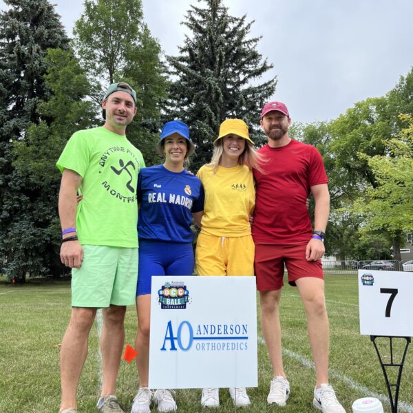 Four teammates pose in front of the Anderson Orthopedics court sponsor sign. The group is each wearing a different neon outfit including green, blue, yellow and red.